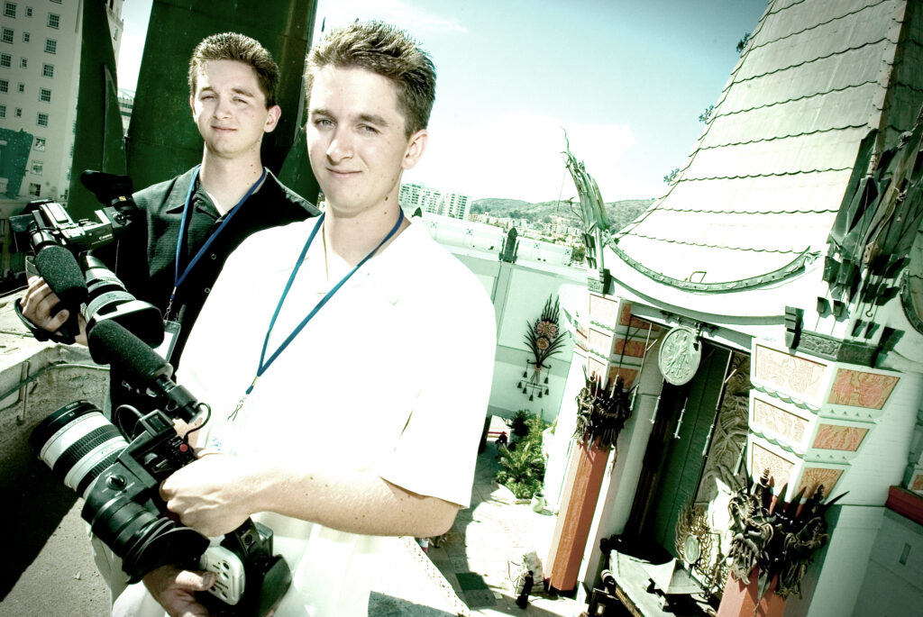 Two men stand together in a bright indoor space as a camera and photographer appear at the left edge.