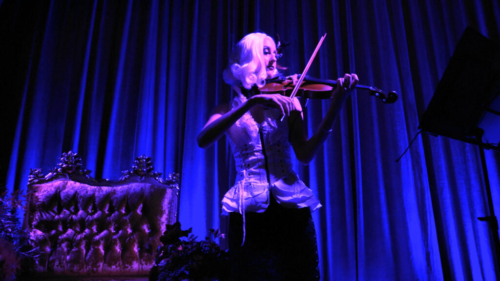A violinist performs beside an ornate chair on a blue-lit stage at the Google Holiday Party.