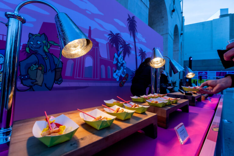 A colorful buffet display with plated dishes sits beneath hanging lamps in a purple-lit event space.