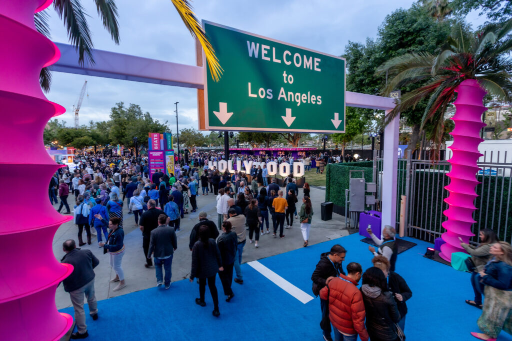 A crowded event entrance beneath a green Welcome to Los Angeles freeway sign, with colorful columns along the walkway.