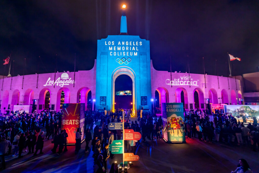 A crowd enters the brightly illuminated Los Angeles Memorial Coliseum beneath the venue's sign and Olympic rings.