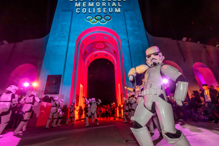 The Los Angeles Memorial Coliseum entrance glows blue and magenta as crowds gather outside for an evening event.