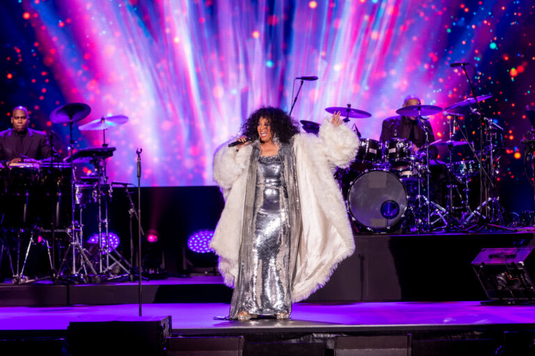 A vocalist in a shimmering outfit performs with a live band against vivid purple and blue stage lights.