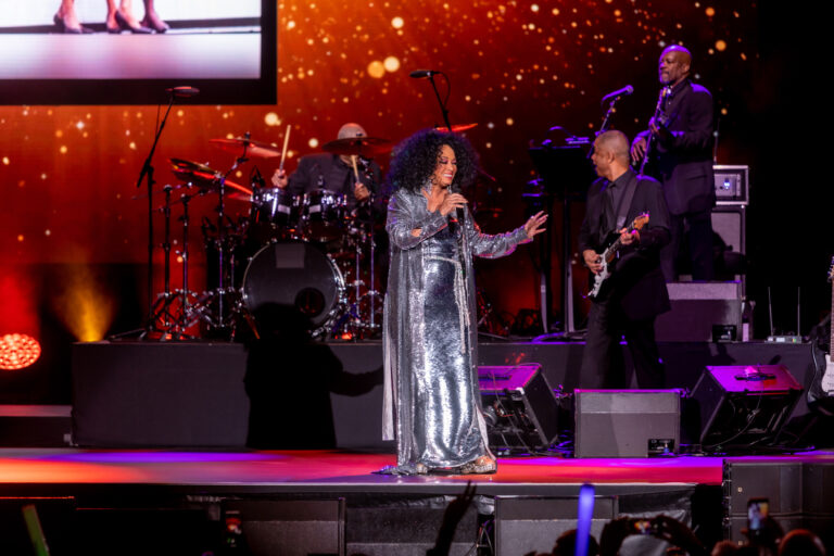 A vocalist in a shimmering outfit performs with a live band under red and purple stage lights.