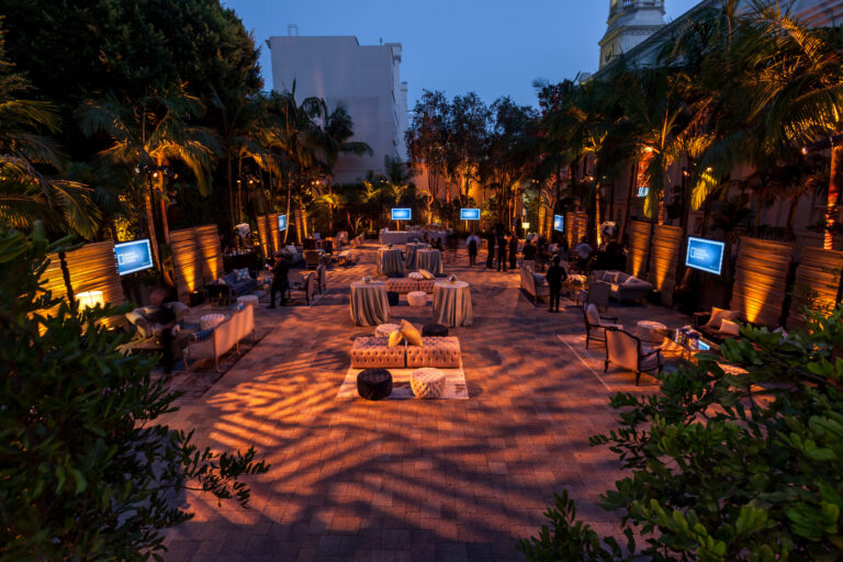 An outdoor event courtyard at dusk is lined with lounge seating, cocktail tables and display screens beneath string lights.