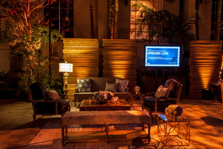 A landscaped outdoor lounge has sofas, armchairs and low tables arranged on a blue rug beneath trees.