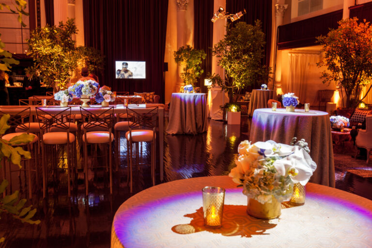 A warmly lit lounge has a central bar, sofas and floral displays arranged beneath arched ceilings.