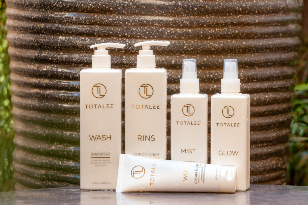 Totallee skincare and hair-care bottles labeled Wash, Rins, Mist and Glow are arranged with a cream elixir tube.