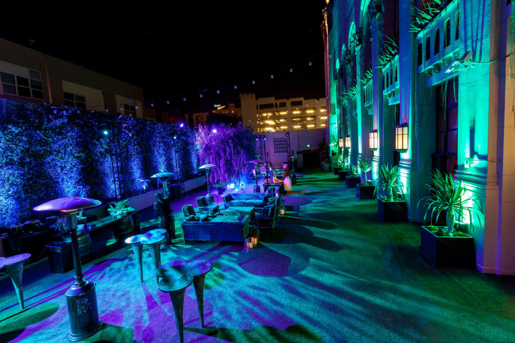 Tall cocktail tables line a blue- and teal-lit reception area beside decorative trees.
