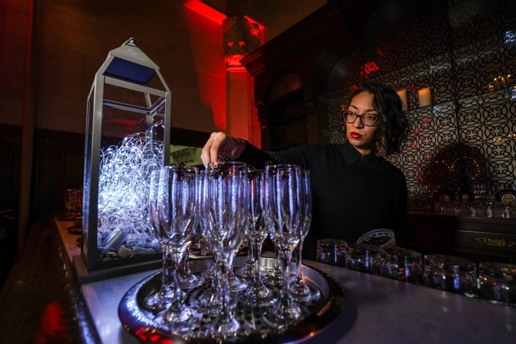 A woman in glasses reaches over rows of champagne flutes at a dimly lit bar.