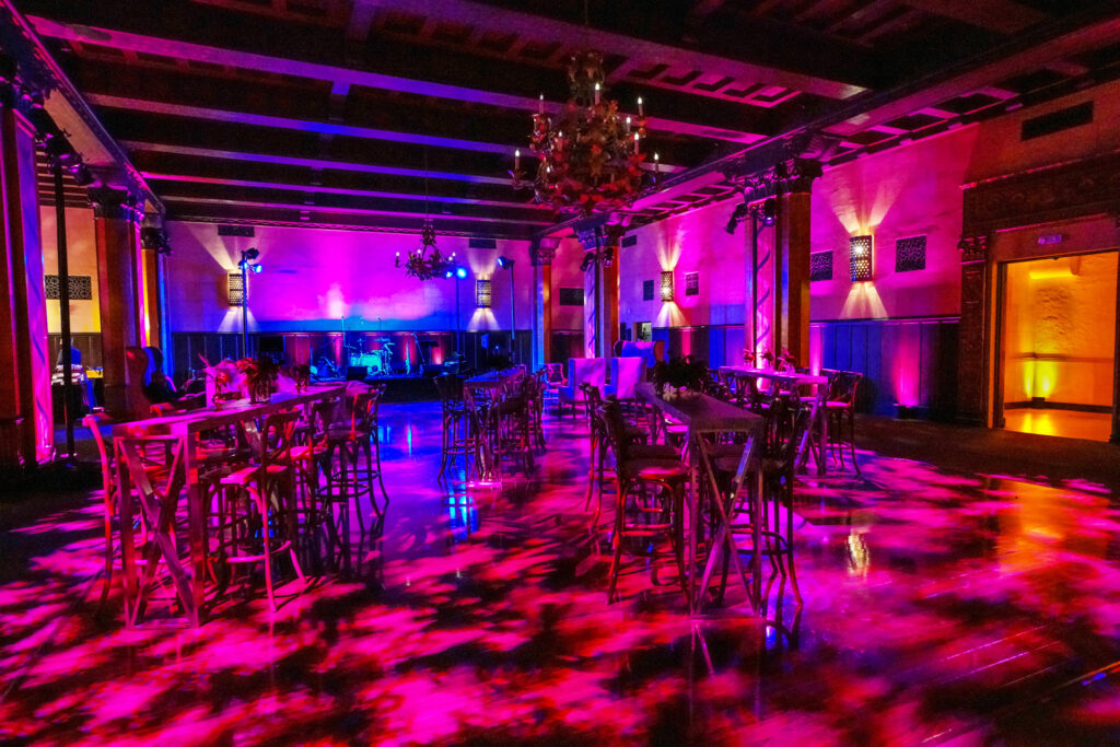 Long tables and rows of chairs fill a warmly lit event room washed in magenta and blue light.