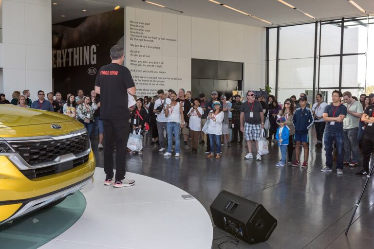 Kia America staff member in a STAGE CREW shirt addressing attendees beside a yellow Kia Soul on display under a GIVE IT EVERYTHING wall with the KIA logo, photographed in Los Angeles.