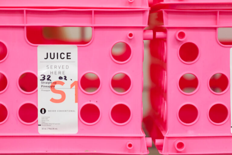 A white juice product card sits in a vivid pink organizer with rows of circular openings.