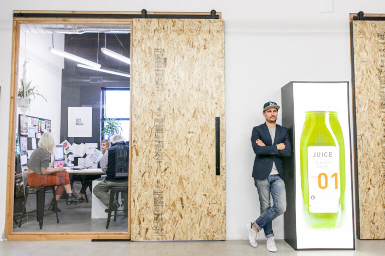 A man in a dark blazer and baseball cap poses beside an oversized juice bottle display in a bright office.