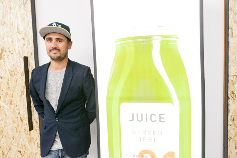 A man in a dark blazer and baseball cap stands beside an oversized green juice bottle graphic.
