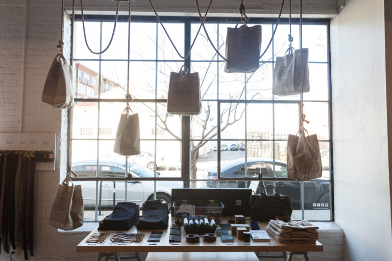 Canvas totes hang from cords above a display table of belts, wallets, folded clothing and small goods.