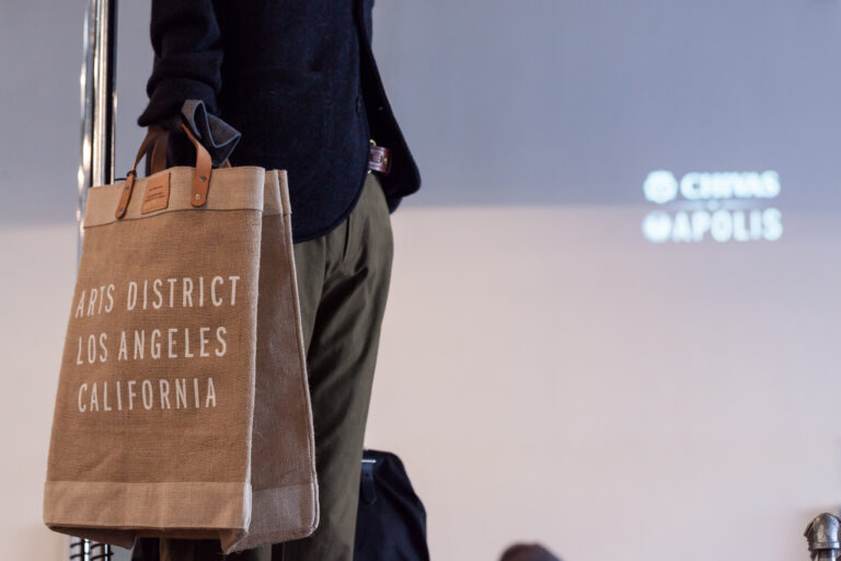 A person holds a tan tote printed “Arts District Los Angeles California” beside a projected Apolis logo.