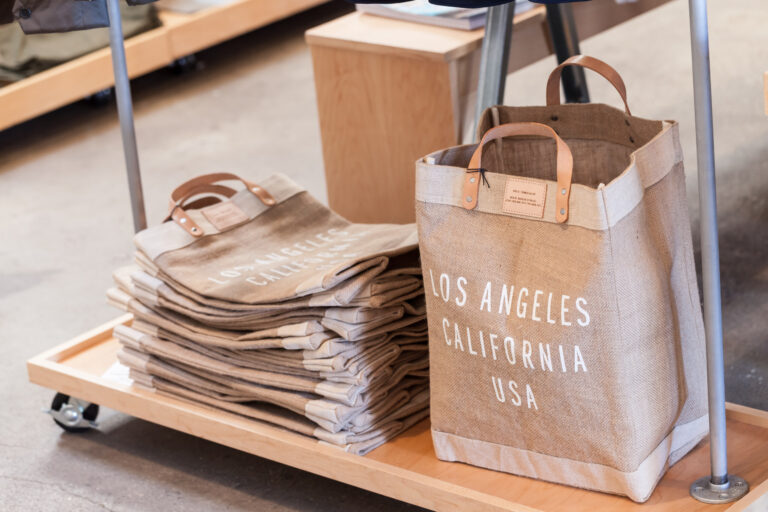 Canvas tote printed “Los Angeles California USA” stands beside a neat stack of folded tan bags on a wooden display.