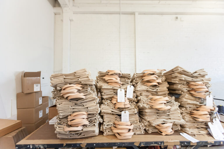 Rows of folded tan canvas tote bags with leather-colored straps and paper tags stacked on a table.