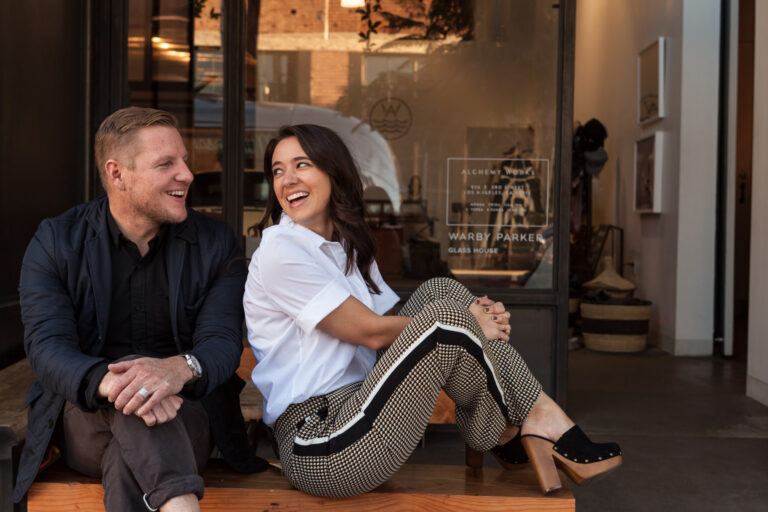 A man and woman sit together on a low step beside a glass storefront entrance.