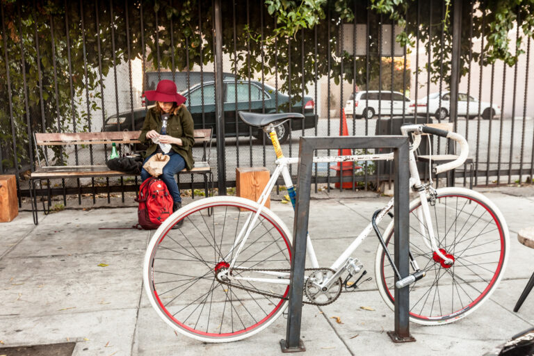 A white bicycle with a red-rimmed front wheel stands outside a storefront as people talk nearby.
