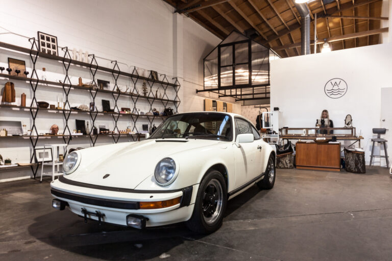 A white classic sports car is parked inside a bright showroom with shelving and retail displays.