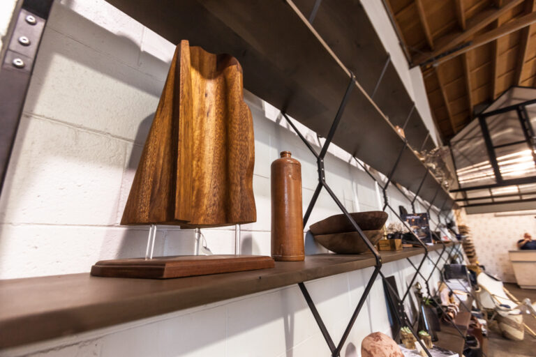 Long carved wood forms and small wooden objects displayed along a wall of dark metal shelves.