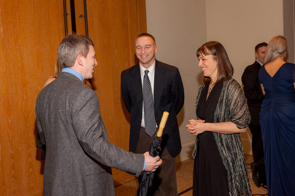 Man shaking hands with another attendee while a woman looks on during a formal professional event.