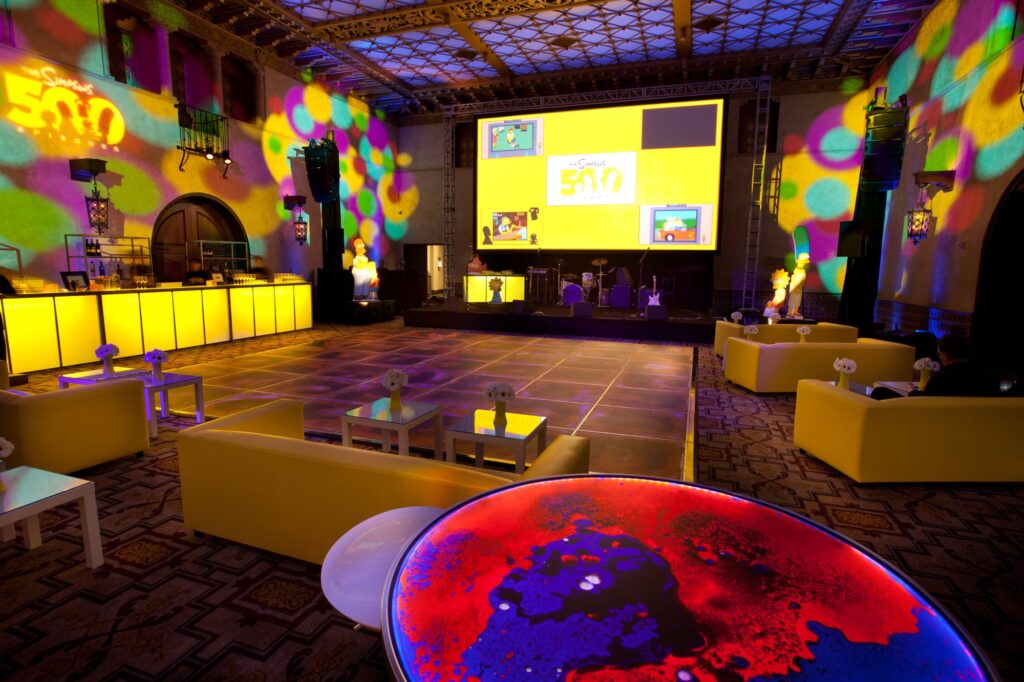 Yellow circular bar and lounge seating beneath a Simpsons screen and colorful event lighting.